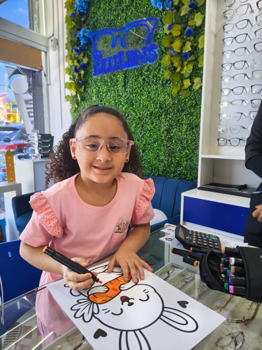 Criança sorrindo usando óculos de grau dentro da Ótica Blulens, segurando um desenho